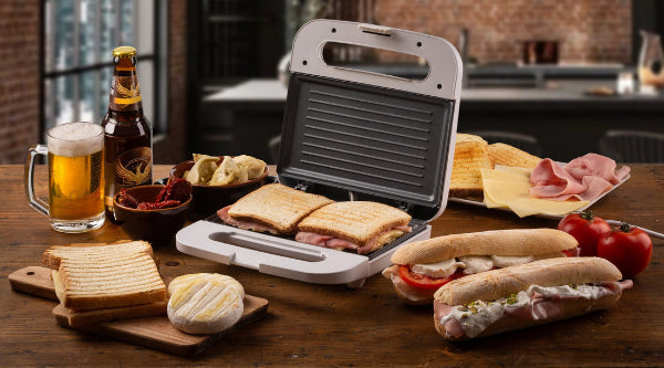 Sandwich maker with bread, meats, cheeses, and a beer on a wooden table.