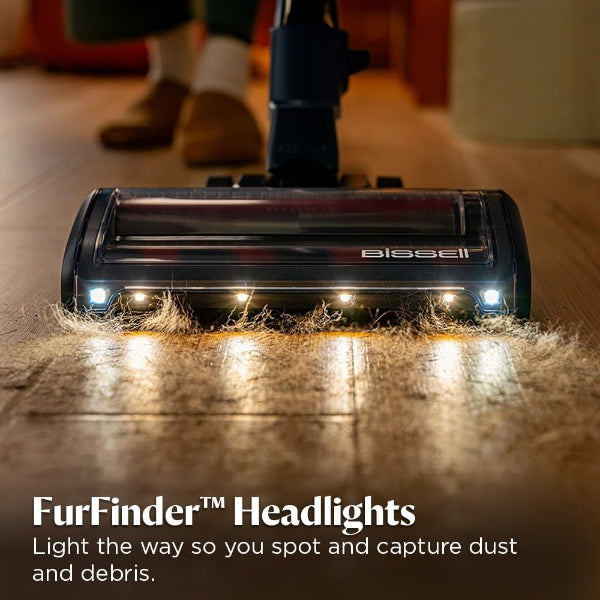 A close-up of a Bissell FurFinder vacuum headlight in action on a wooden floor, illuminating dust and debris with bright LED lights. The text at the bottom reads 'FurFinder™ Headlights' and 'Light the way so you spot and capture dust and debris.