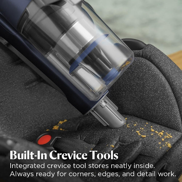Close-up of a dark blue and silver crevice vacuum cleaner being used to clean the fabric of a dark gray upholstered chair, with yellow dust particles visible on the surface. White text at the bottom reads 'Built-In Crevice Tools' and 'Integrated crevice tool stores neatly inside.