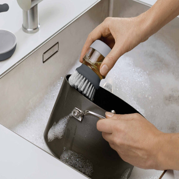 A person using the Joseph Joseph Palm Scrub™ Grey Washing-Up Brush to scrub a pot in the sink, with soap dispensing mechanism in use.